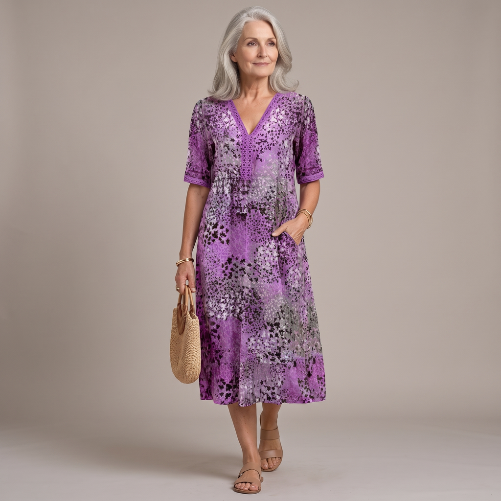 An older woman stands against a plain beige studio background wearing a purple floral midi dress with short sleeves, a V-neckline, and side pockets. She is holding a woven straw tote bag and wearing beige sandals.