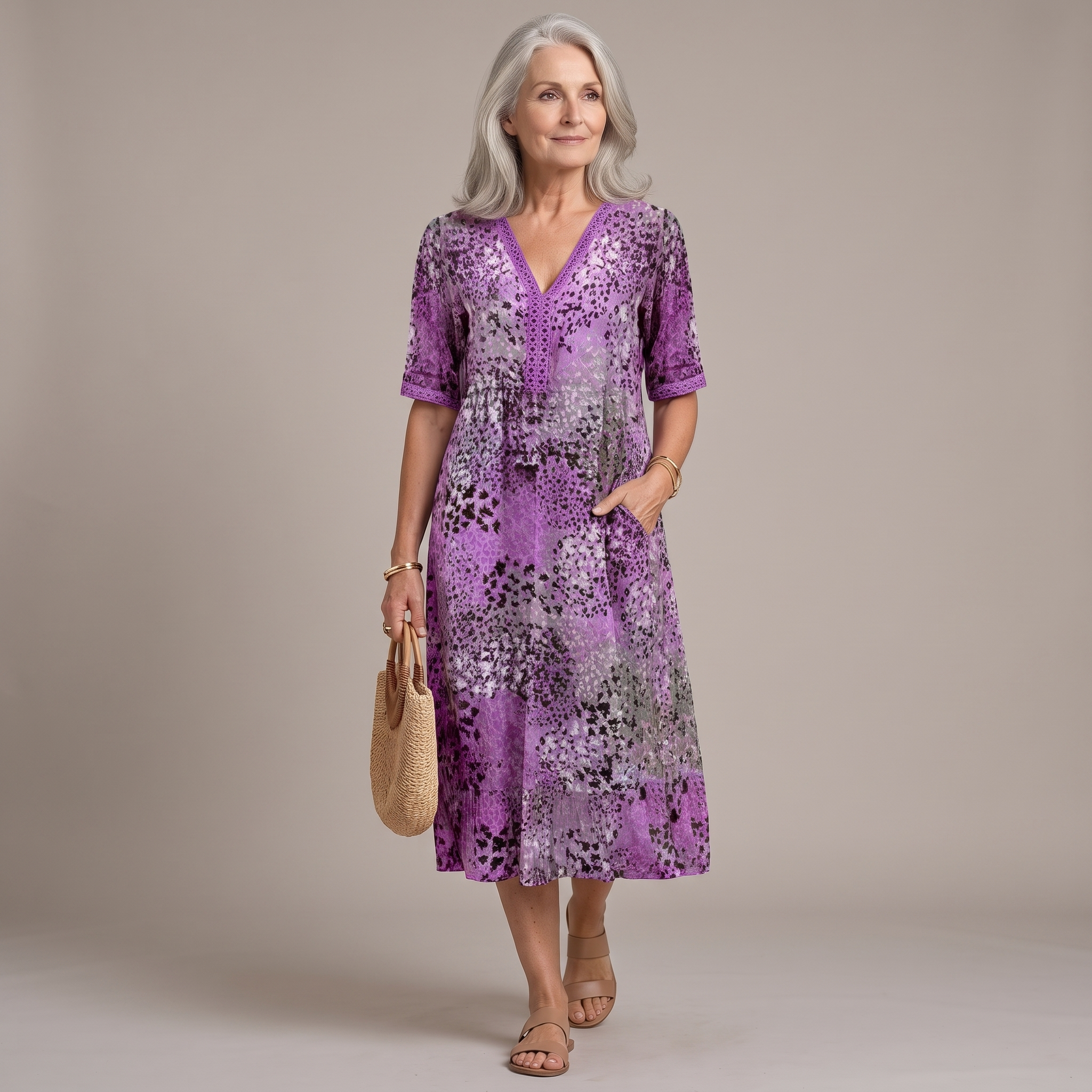 An older woman stands against a plain beige studio background wearing a purple floral midi dress with short sleeves, a V-neckline, and side pockets. She is holding a woven straw tote bag and wearing beige sandals.
