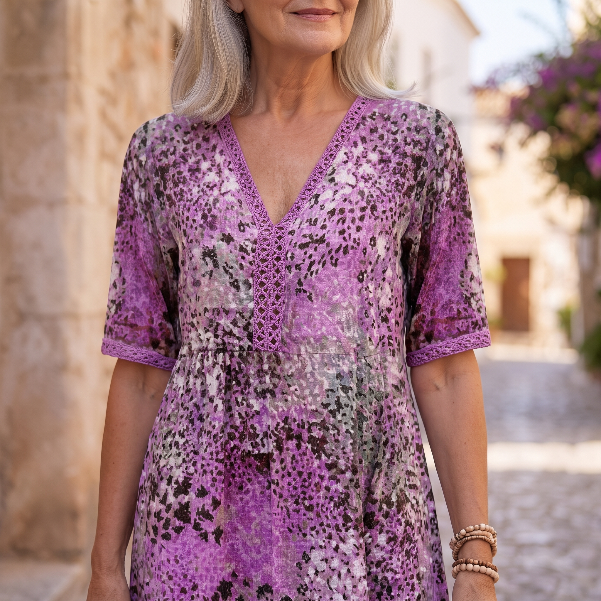 A woman wearing a purple and black leopard-print lace dress with a V-neckline and elbow-length sleeves, photographed outdoors in a sunlit stone-walled setting.