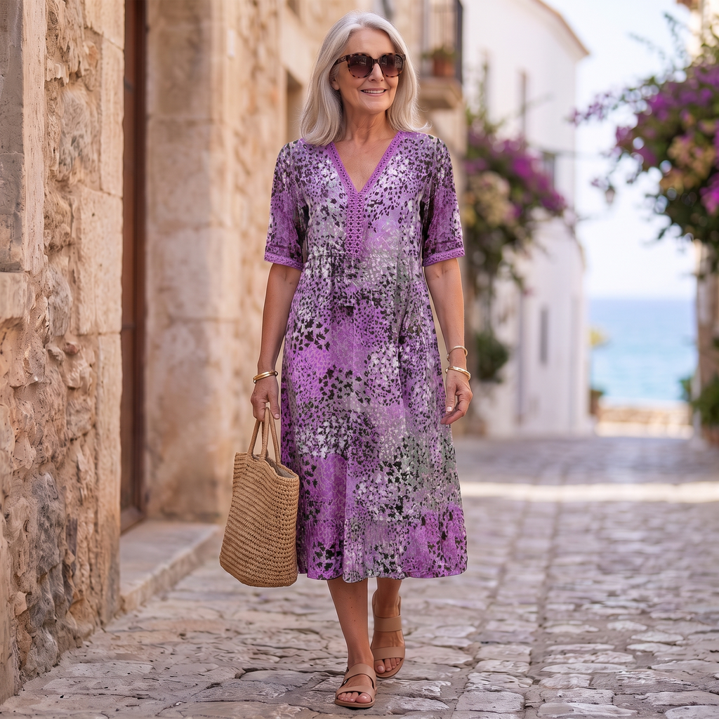 An older woman walking down a cobblestone street wearing a purple short-sleeve midi dress with a black and gray abstract floral pattern, holding a woven straw tote bag.