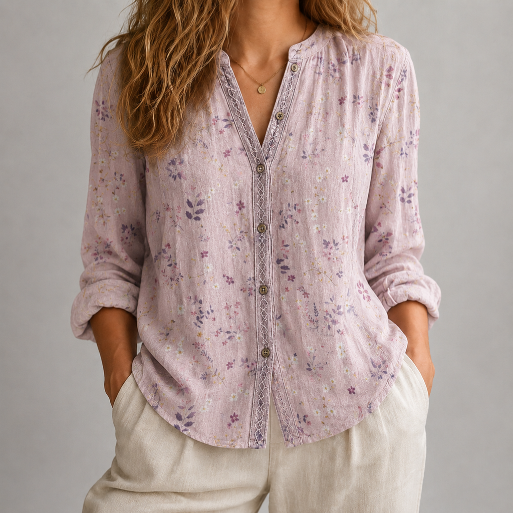 A woman wearing a light pink floral button-up blouse with lace trim and cream pants, photographed against a plain gray studio background.