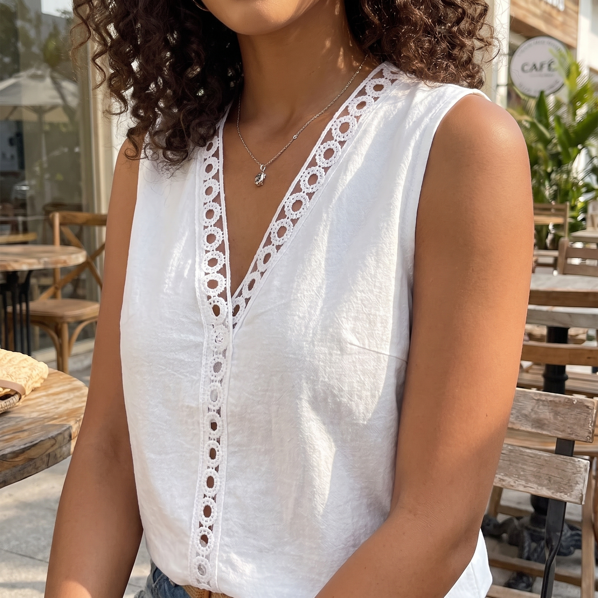 A woman wearing a white sleeveless V-neck top with decorative eyelet lace trim down the front, photographed in an outdoor cafe setting.